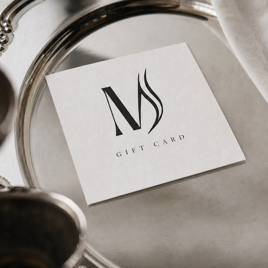 A white square gift card with a black stylized 'M' logo and the words 'GIFT CARD' printed below, placed on an ornate silver tray with a white cloth in the background.