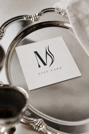 A white square gift card with a black stylized 'M' logo and the words 'GIFT CARD' printed below, placed on an ornate silver tray with a white cloth in the background.